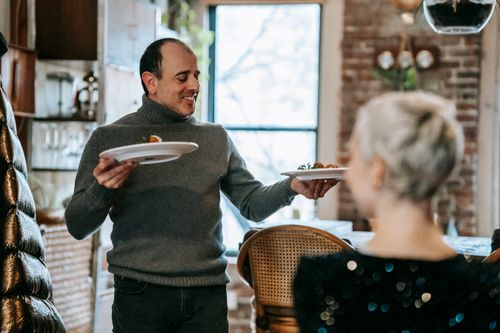 Free Positive middle aged couple having dinner in bright room while man serving plates with tasty food near chairs and counter near window Stock Photo