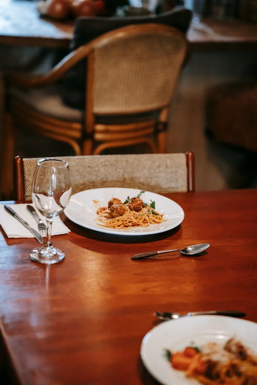 Plates with tasty pasta with meatballs and herbs with tomatoes on wooden table near fork and knife with spoon and empty glass near chair in bright restaurant