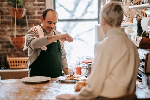 Free Adult couple in light kitchen near counter while male salting pasta on plates near onion and kitchenware near window in daytime while lady sitting nearby Stock Photo