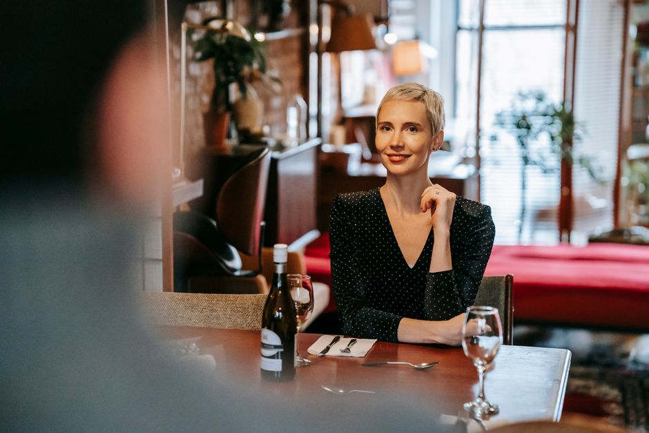 Couple having date in bright room while man standing near table with glasses and wine bottle near cutlery while woman sitting in chair in daytime