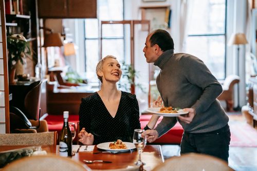Free Adult couple having date in light room near table while male serving plates with pasta with meatballs near cutlery and bottle with wine near glasses while looking at each other Stock Photo