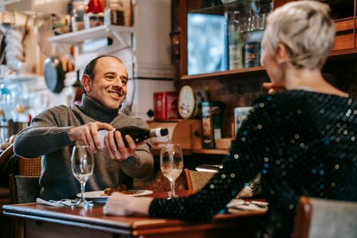 Free Positive couple having date at table while man pouring wine from bottle into wineglasses near plates with tasty dish in bright restaurant while looking at each other Stock Photo