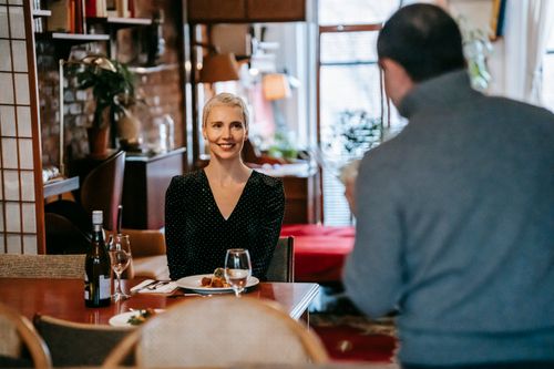 Free Positive adult couple having date in bright room while man standing with bouquet with flowers near chairs and table with glasses and wine bottle near plate with food and cutlery Stock Photo