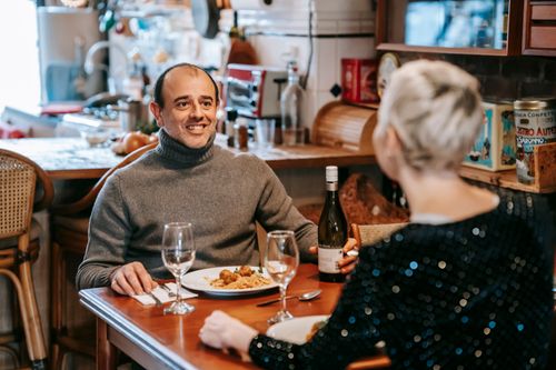 Free Couple sitting at wooden table while having dinner with pasta with meatballs on plates near wineglasses and bottle with alcohol near chairs in light room while looking at each other Stock Photo
