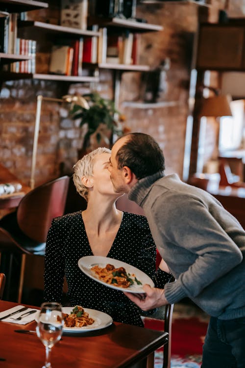 Couple having date while man kissing woman in cheek near table with wineglass near plates with pasta with meatballs near cutlery in light room