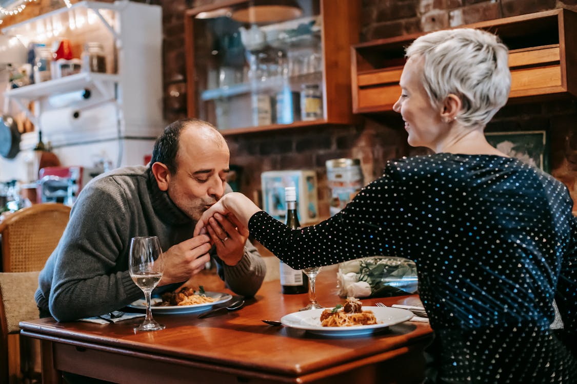 Free Couple having romantic dinner while male kissing female hand while sitting at table with wineglasses and bottle with wine near plate with pasta with meatballs in bright room Stock Photo