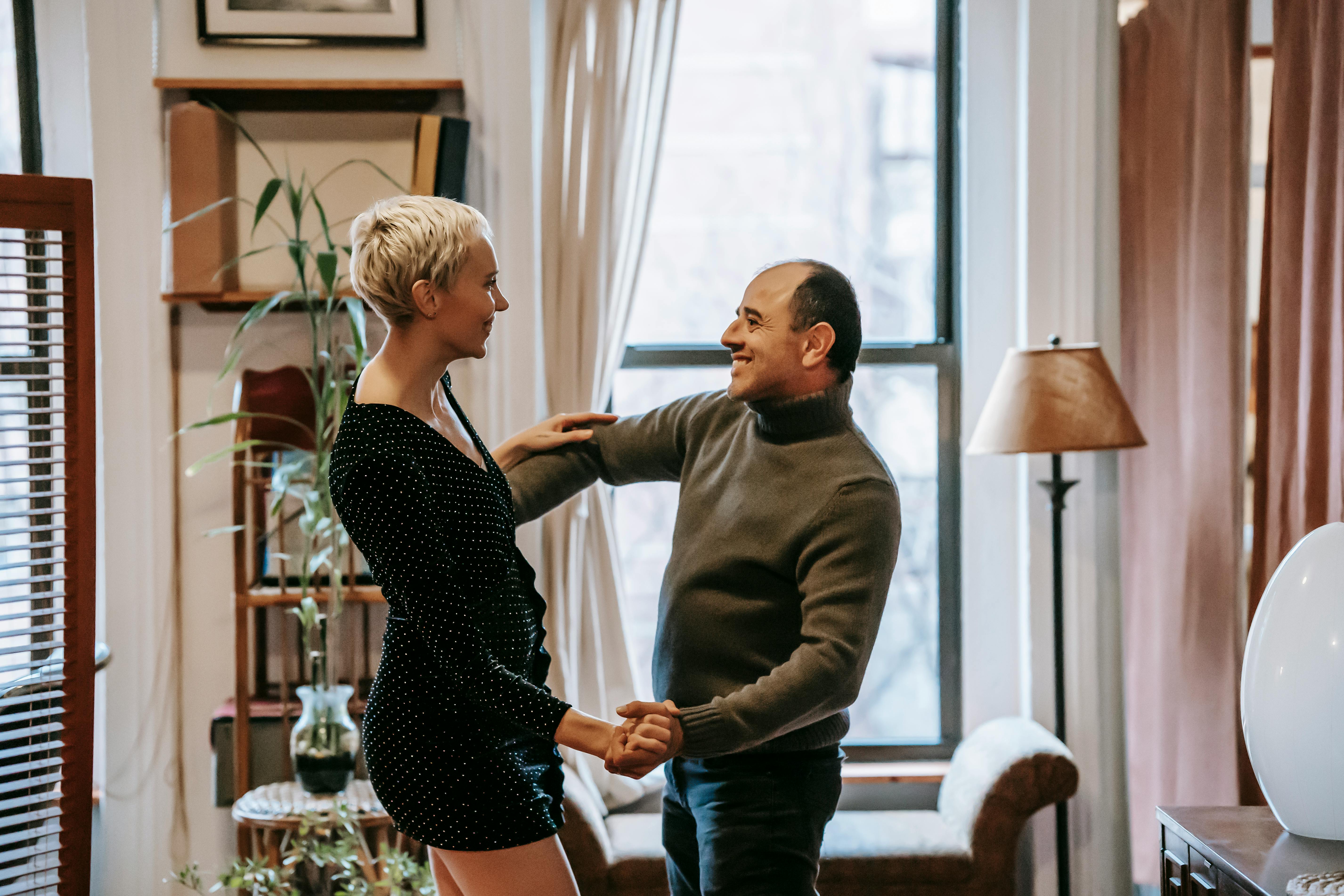 positive couple dancing while holding hands in house near window