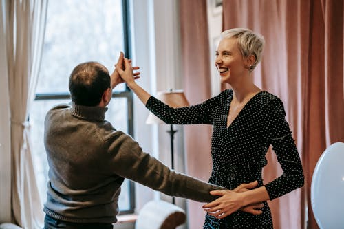 Happy couple holding hands while dancing in room near window