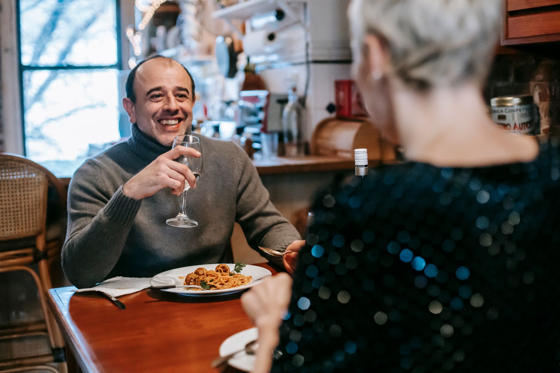 Couple sitting at table while having dinner with pasta with meatballs on plate with glass in light room with chairs near window