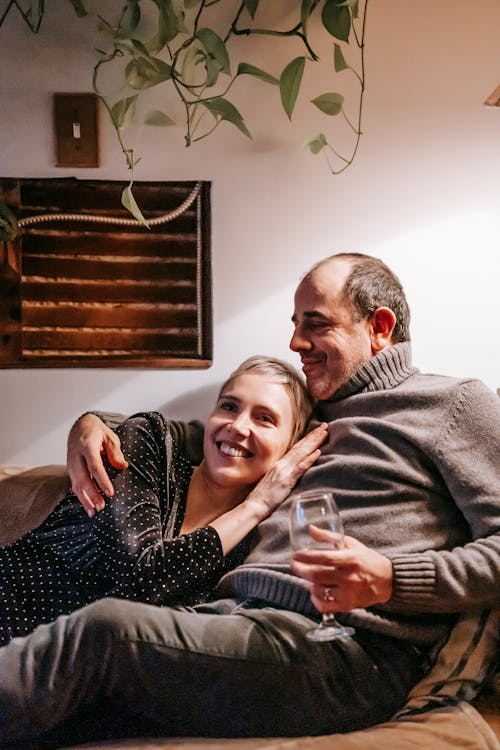 Positive man with glass of drink cuddling smiling wife lying on couch and enjoying pleasant time together