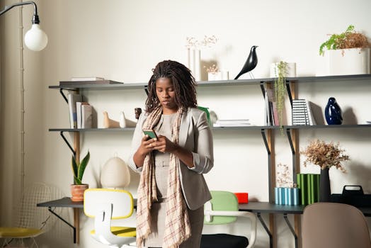 A woman standing in a modern office environment, focused on her smartphone, surrounded by books and plants.