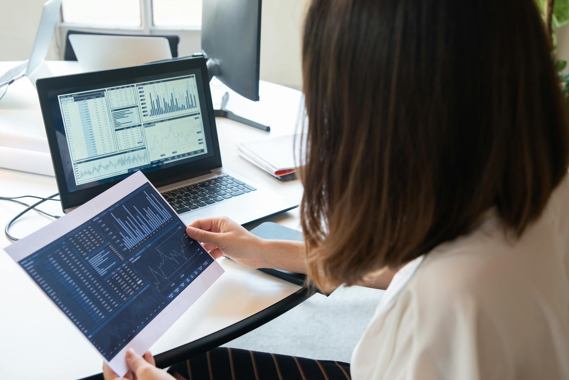 A woman analyzing financial data, holding a printed chart while looking at a laptop displaying various graphs and spreadsheets.