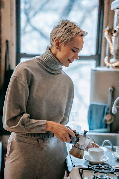 Free Positive female pouring brewed hot beverage from geyser coffee maker into ceramic mug while standing near stove in kitchen Stock Photo