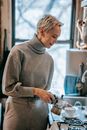 Positive female pouring brewed hot beverage from geyser coffee maker into ceramic mug while standing near stove in kitchen