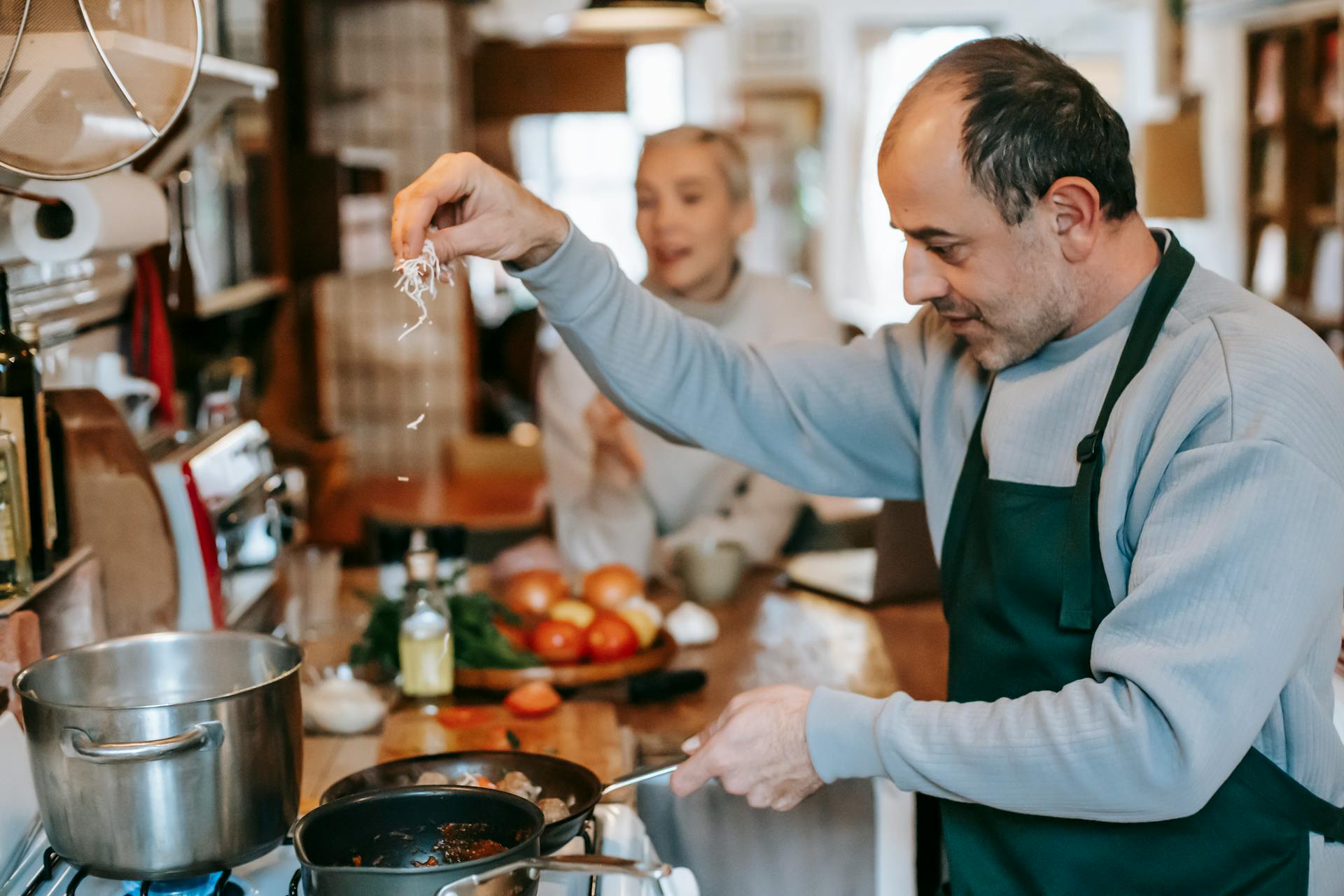 Focused male in apron adding ingredient into frying pan heating on gas stove while cooking meal for woman