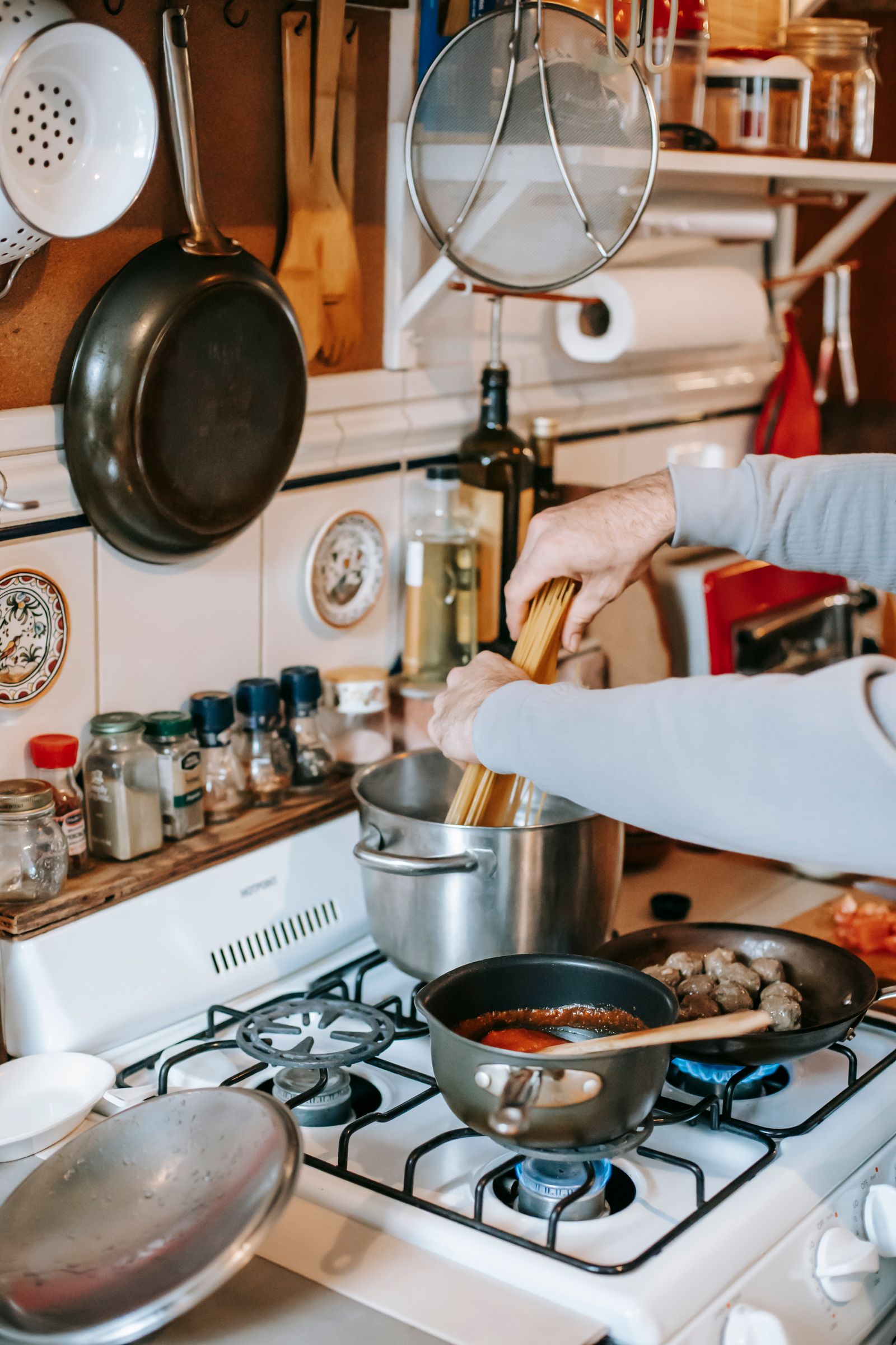 Crop anonymous person putting raw pasta in saucepan on gas stove with ingredients in frying pan and ladle