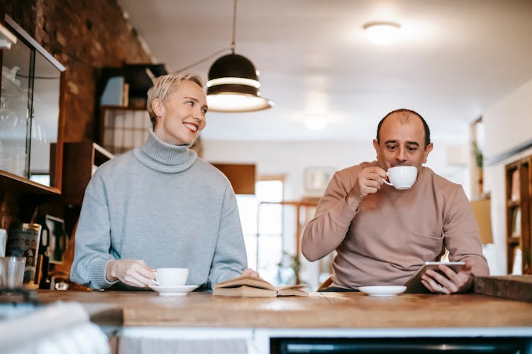 Positive Couple Drinking Coffee In Cafe With Book And Tablet