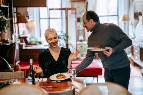 Free Positive adult couple having date and man serving plates with pasta for woman at table with bottle near wineglasses and cutlery Stock Photo