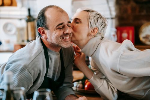 Free Positive adult couple in casual clothes having date while woman kissing man in cheek near counter in light kitchen with products and kitchenware Stock Photo