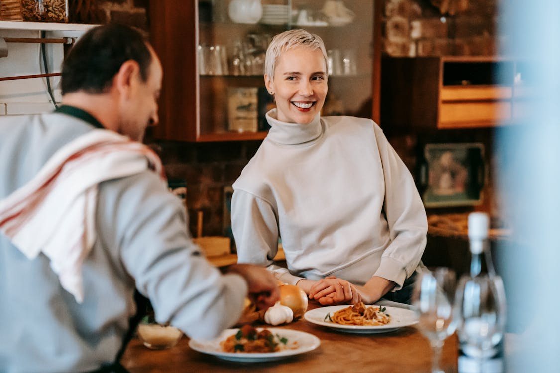 Free Smiling adult couple in casual clothes having dinner while male cooking pasta on plates at counter near glasses and wine bottle near various products and ingredients in light kitchen Stock Photo