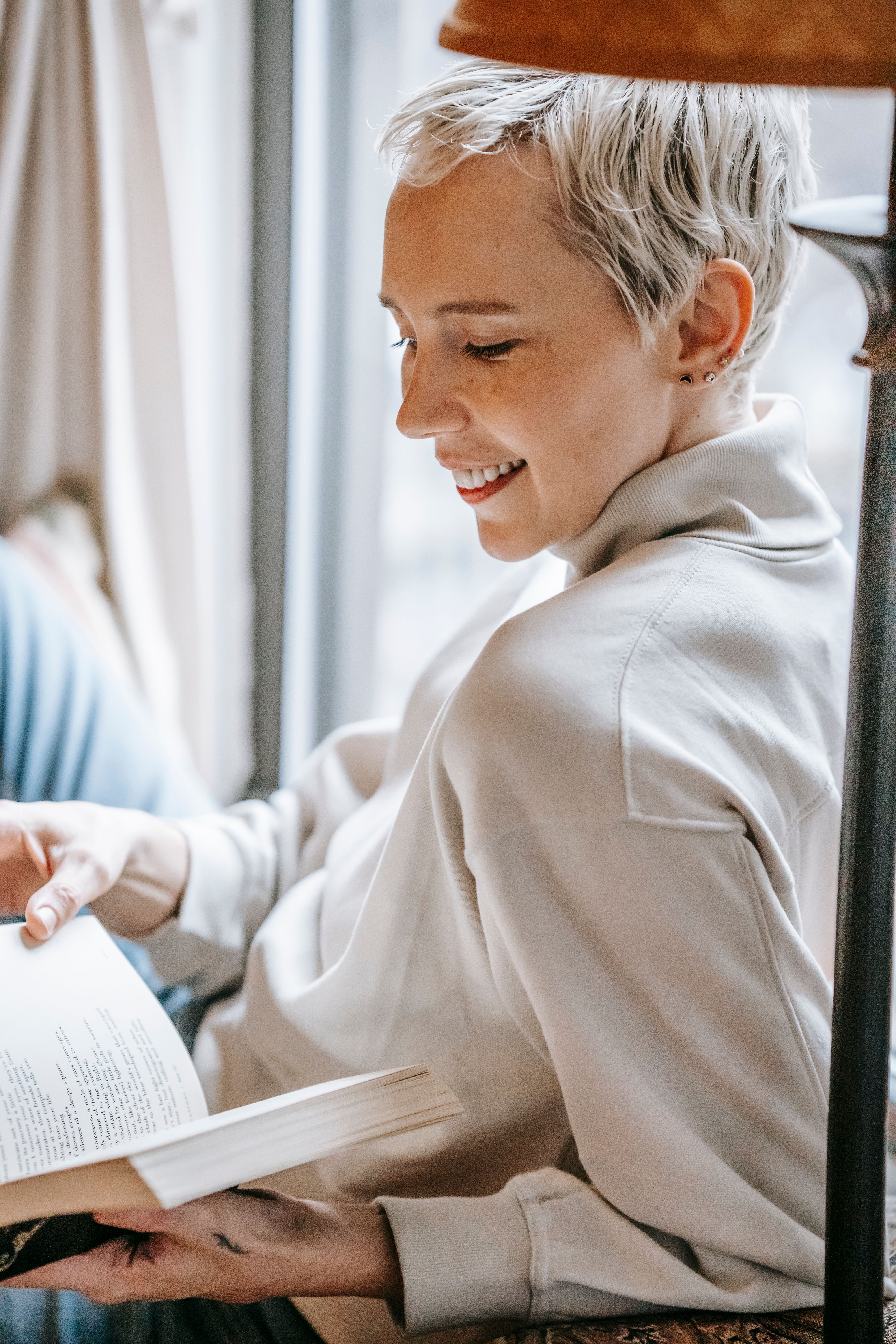 Side view of positive female smiling widely while reading book and enjoying leisure lying against window. pexels, side, view