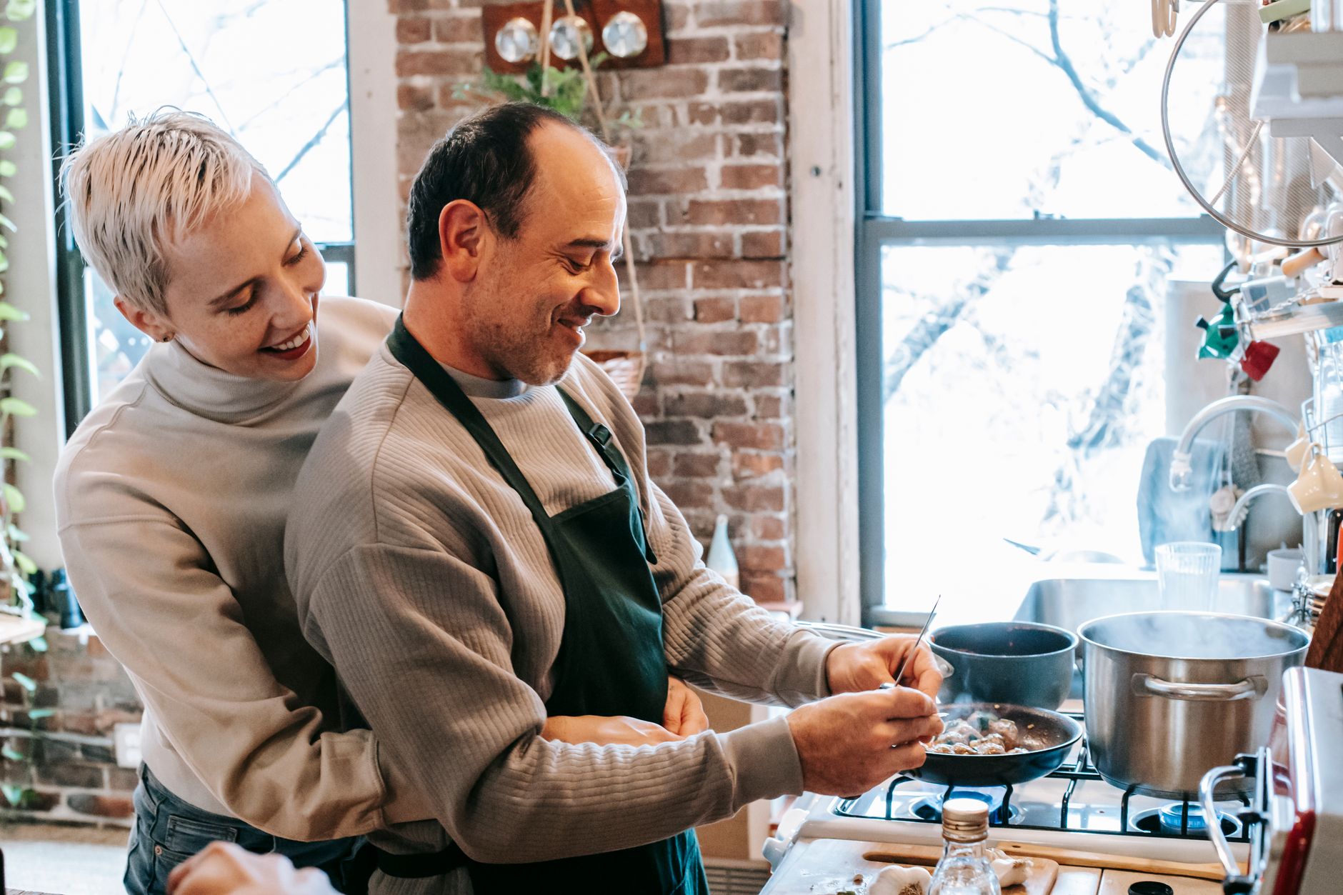 Side view of gentle cheerful wife hugging husband in apron standing at table and peeling garlic while cooking lunch