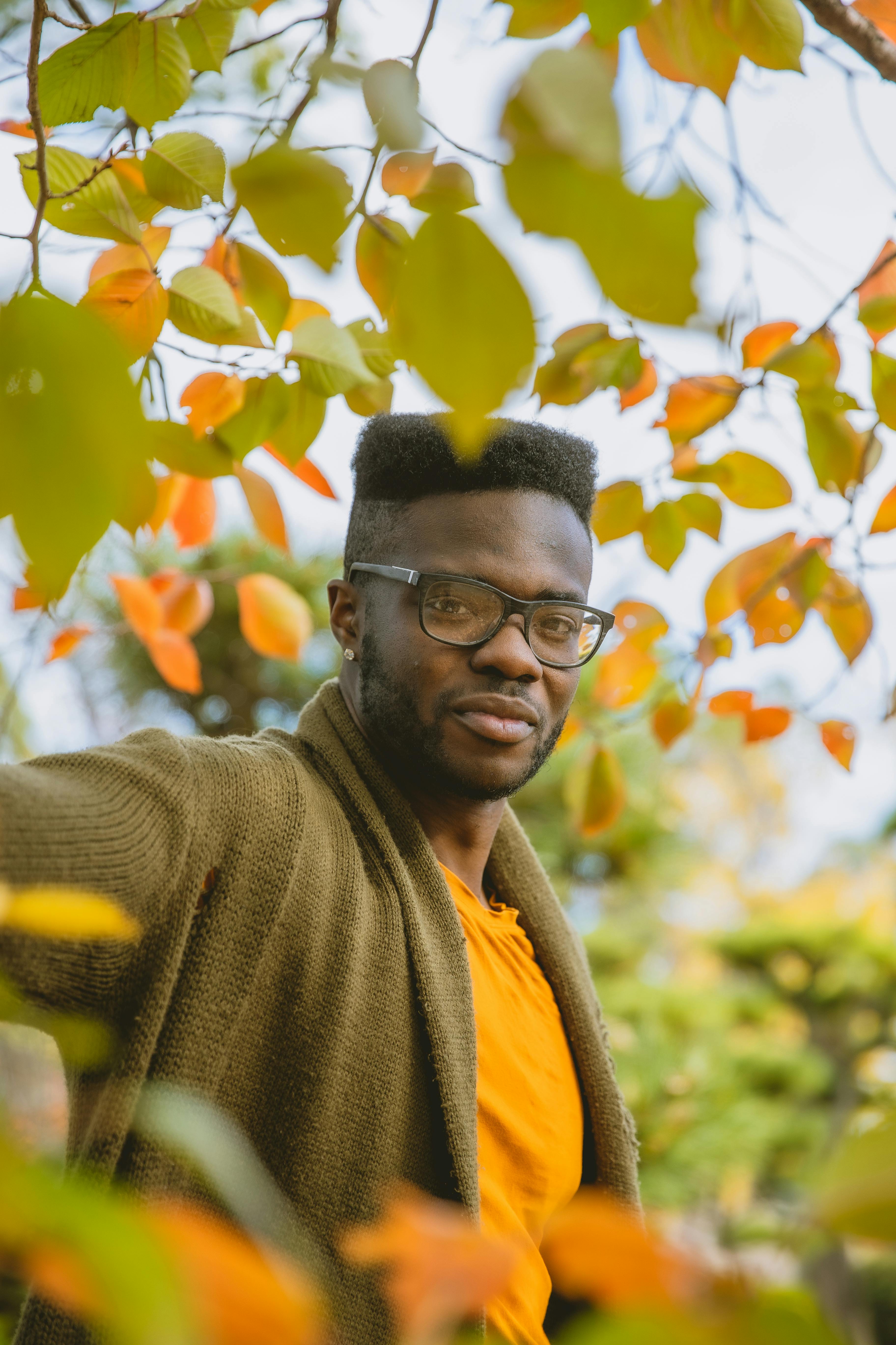 Positive young African American male in casual outfit and eyeglasses resting in autumn park near trees and plants with bright yellow leaves and looking at camera