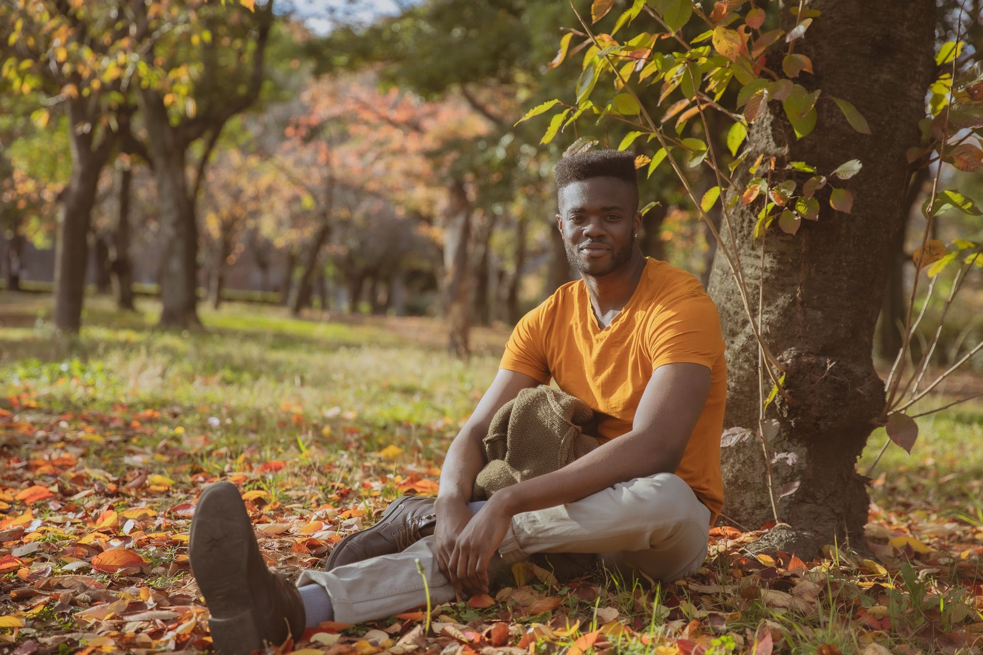 Full body of young happy African American male in casual outfit smiling and looking at camera while relaxing on grassy lawn covered with fallen leaves in autumn park