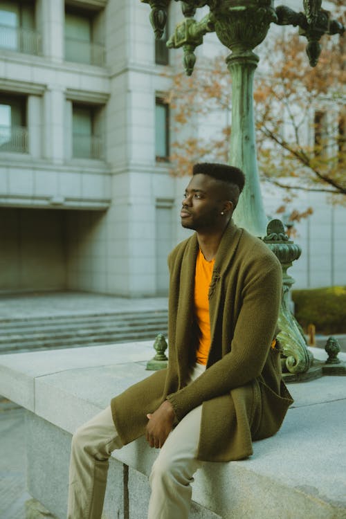 Side view of pensive African American male in trendy outfit sitting on concrete surface near pillar on street near building in city