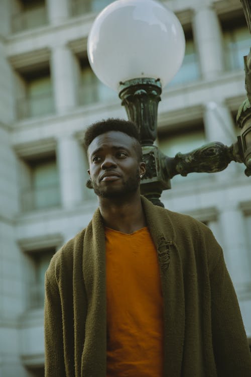 Pensive African American male in stylish outfit looking away while standing near streetlight in city against residential building on blurred background