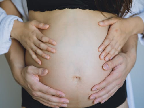 Unrecognizable pregnant wife in black underwear and anonymous gentle husband touching bare belly with hands while standing in room near wall