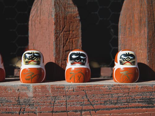 Collection of red painted traditional Japanese daruma dolls placed on shabby wooden plank under sunlight on street in suburb area