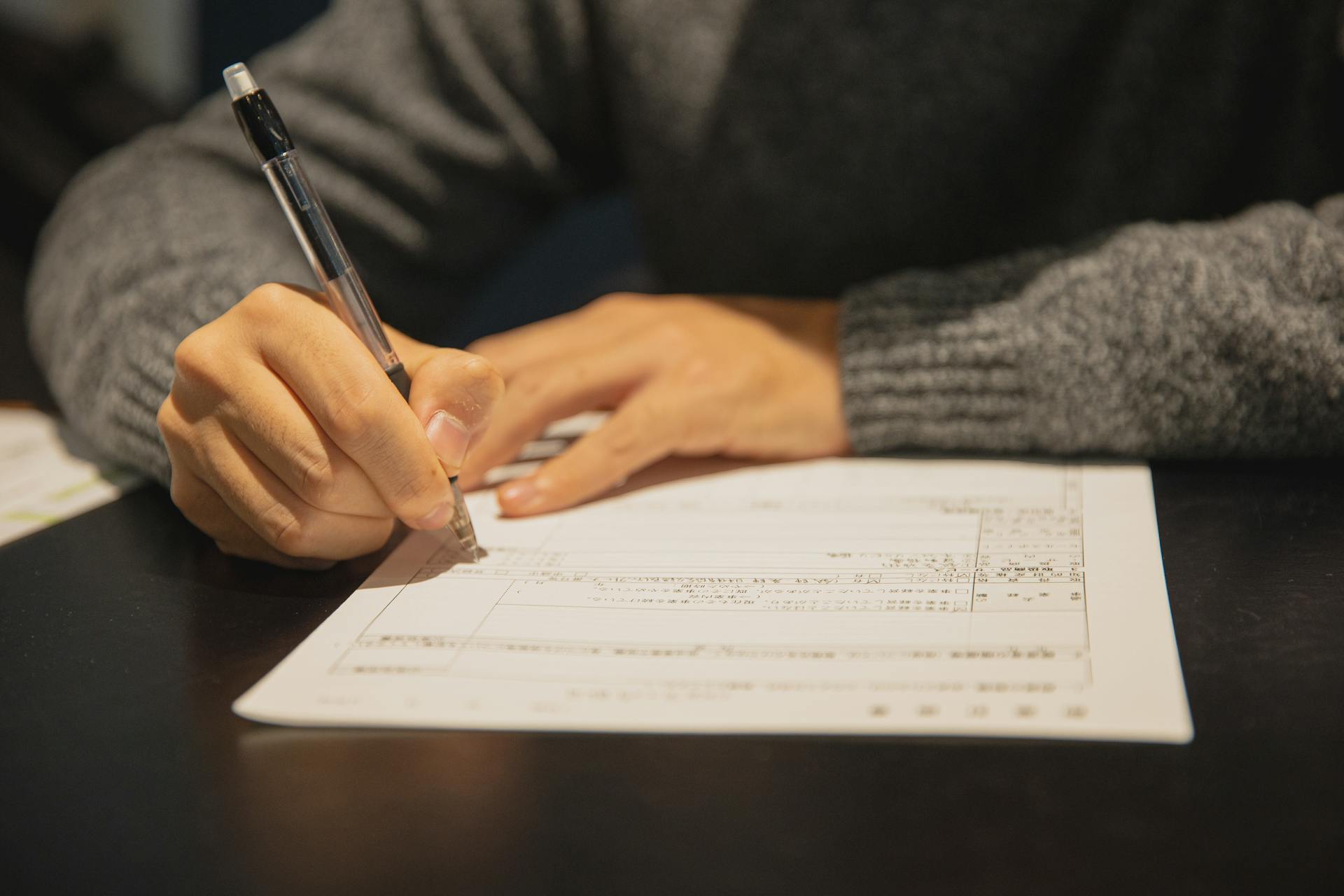 Close-up of a man in a gray sweater signing a document at a desk in an office setting.