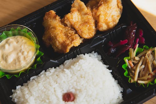 From above of plastic container with fried chicken and rice with sauce near sauce and sliced vegetables