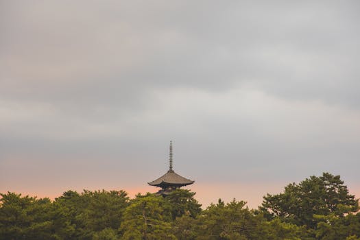 A tranquil view of a traditional Japanese pagoda surrounded by lush green trees at sunset under a cloudy sky.