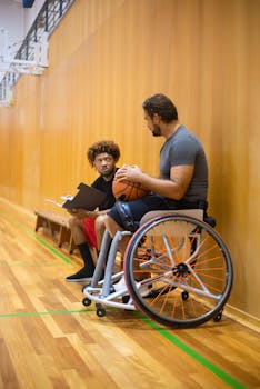 Man in wheelchair discussing with coach in a gym, emphasizing inclusive sports.