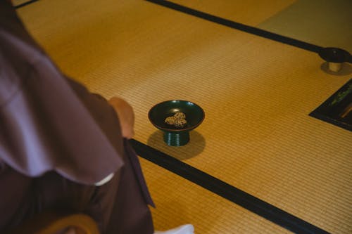Side view of unrecognizable person in traditional robe sitting in tea room with tenmoku tea bowl placed on tatami during traditional Japanese ceremony