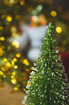 A close-up of a glittery miniature Christmas tree with festive bokeh lights in the background.