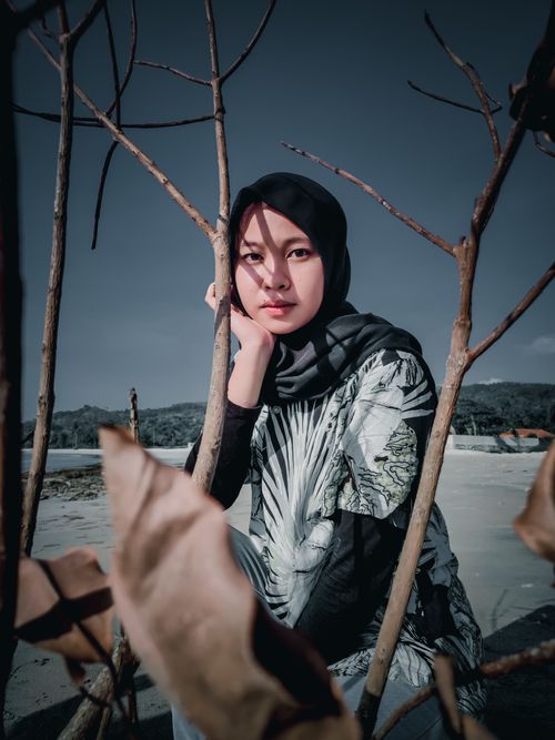Free A woman in a hijab posing thoughtfully by the beach with dry branches in foreground. Stock Photo