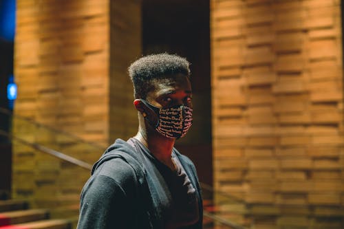 Free Calm African American male wearing medical mask looking at camera while standing on street near building and staircase in evening time Stock Photo