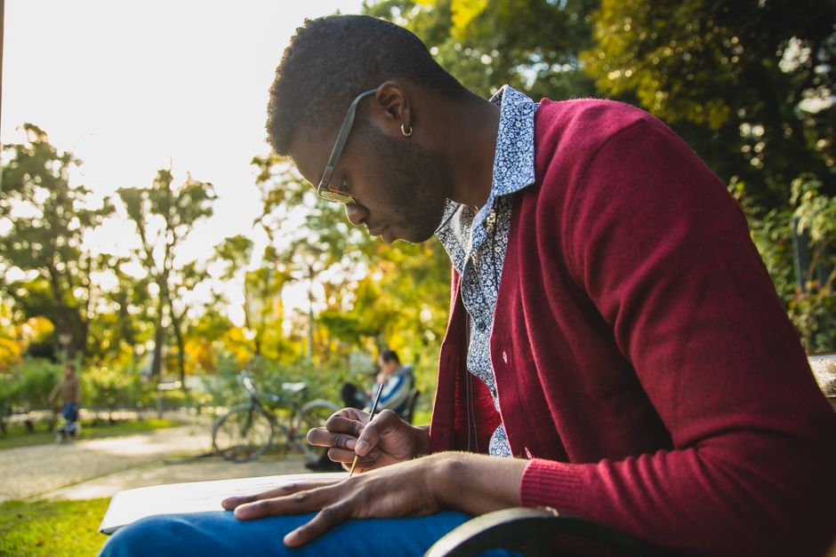 Side view of young African American man in eyeglasses taking notes on paper while doing homework outdoors Side view of young African American man in eyeglasses taking notes on paper while doing homework outdoors