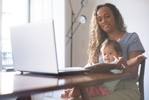 Free A mother multitasks at a desk with a laptop and her child in a bright home setting. Stock Photo