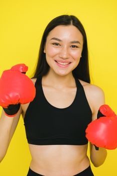 Confident woman in black sports bra and red boxing gloves smiles brightly against a vibrant yellow backdrop.