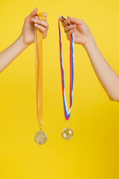 Two hands holding gold medals with colorful ribbons on a bright yellow background.