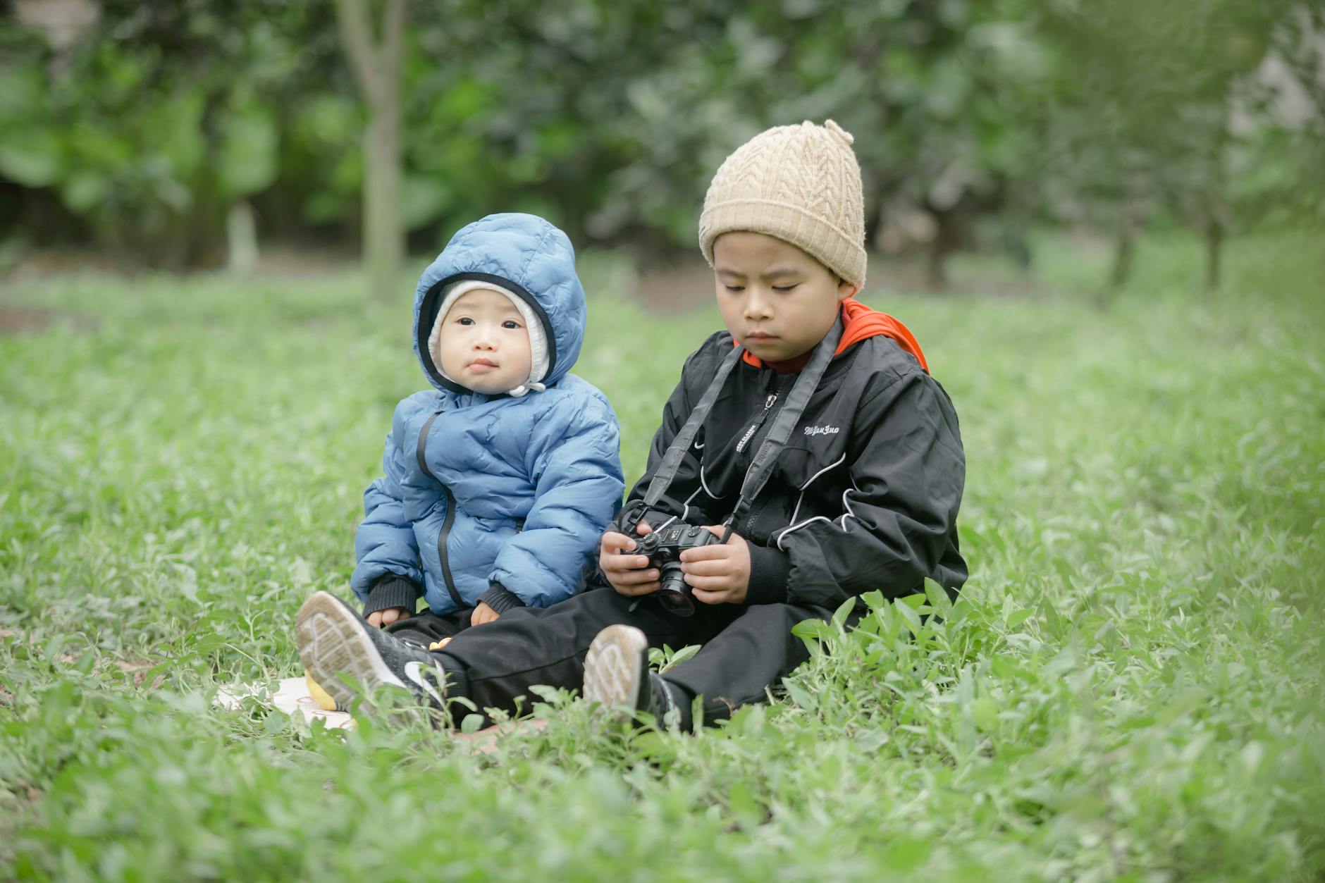 Two children sitting on grass in winter clothes, one holding a camera, in a tranquil outdoor setting.