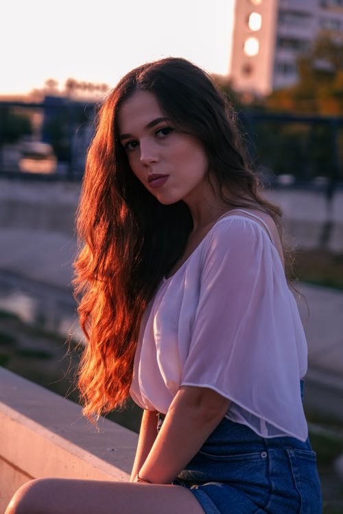 Free Portrait of a woman with long hair in denim shorts sitting outdoors at sunset. Stock Photo