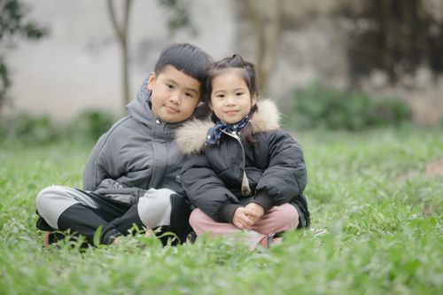 Free Two Asian children in warm coats sitting on grass, embracing the winter outdoors. Stock Photo