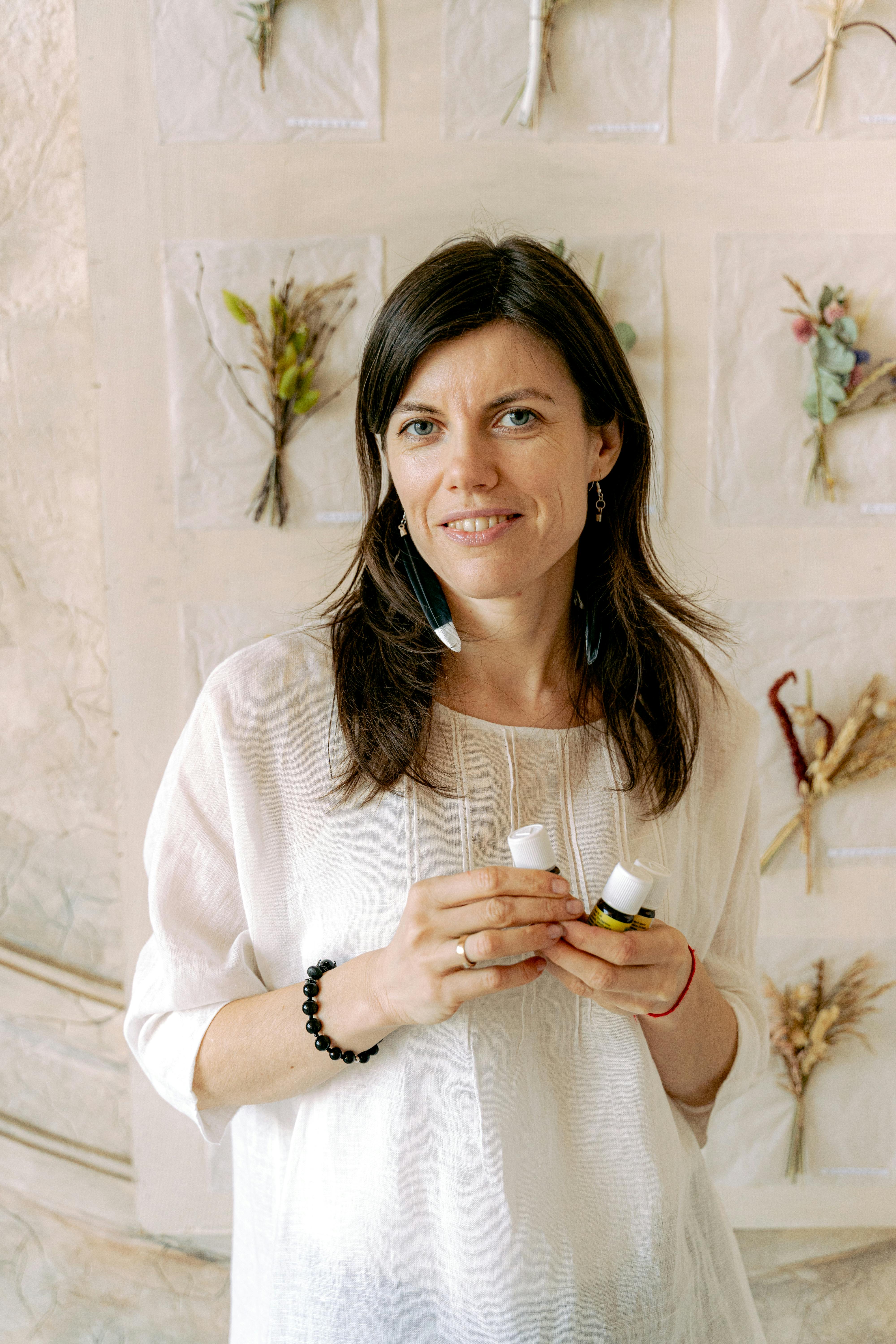 A woman in a white blouse, holding skincare products, smiles at the camera indoors.