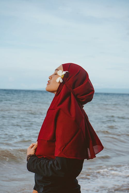 Side view of peaceful Muslim female wearing hijab standing on beach with closed eyes and enjoying fresh breeze