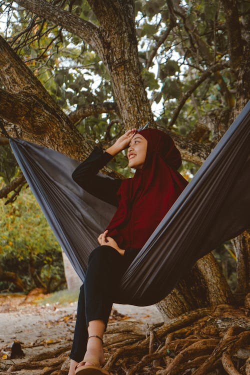 From below of smiling Muslim woman sitting in hammock with crossed legs during trip in exotic country